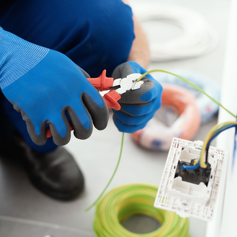 Électricien en gants bleus coupant un fil vert-jaune avec une pince rouge près d'une prise murale nue et de bobines de câble.