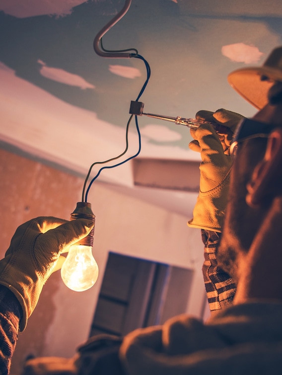 Électricien avec gants tenant une ampoule allumée tout en testant un fil électrique avec un tournevis testeur au plafond.
