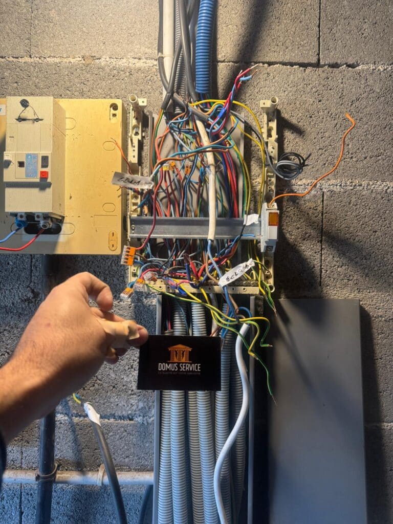 Problème câblage tableau électrique - Intervention électricien Main tenant une carte de visite DOMUS SERVICE devant un tableau électrique résidentiel désordonné avec fils dénudés et étiquettes manuscrites.